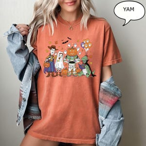 Puede incluir: Camiseta naranja con un gr&aacute;fico de dibujos animados de Halloween. El dise&ntilde;o incluye personajes disfrazados de Woody, un fantasma, un robot con cabeza de calabaza y un dinosaurio. Un bocadillo dice "YAM".