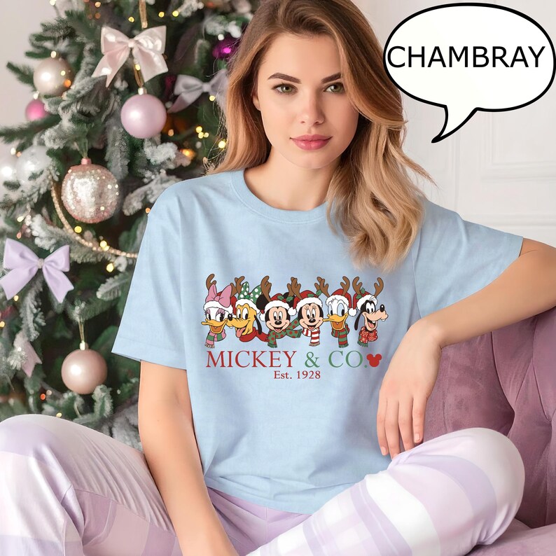Puede incluir: Camiseta azul claro con un dise&ntilde;o festivo de personajes de Disney con cuernos de reno y gorros de Pap&aacute; Noel. El texto "Mickey & Co. Est. 1928" est&aacute; en rojo. La persona lleva un pantal&oacute;n de pijama lila claro.