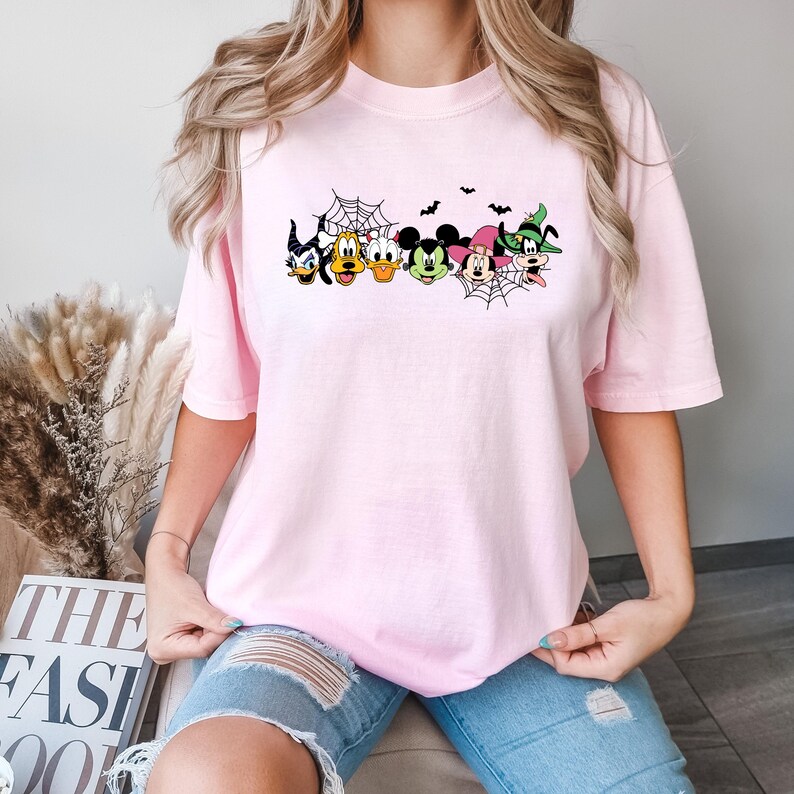 May include: A pink t-shirt featuring a graphic of Mickey Mouse, Donald Duck, Goofy, and other Disney characters dressed in Halloween costumes. The characters are arranged in a line with a spiderweb behind them.