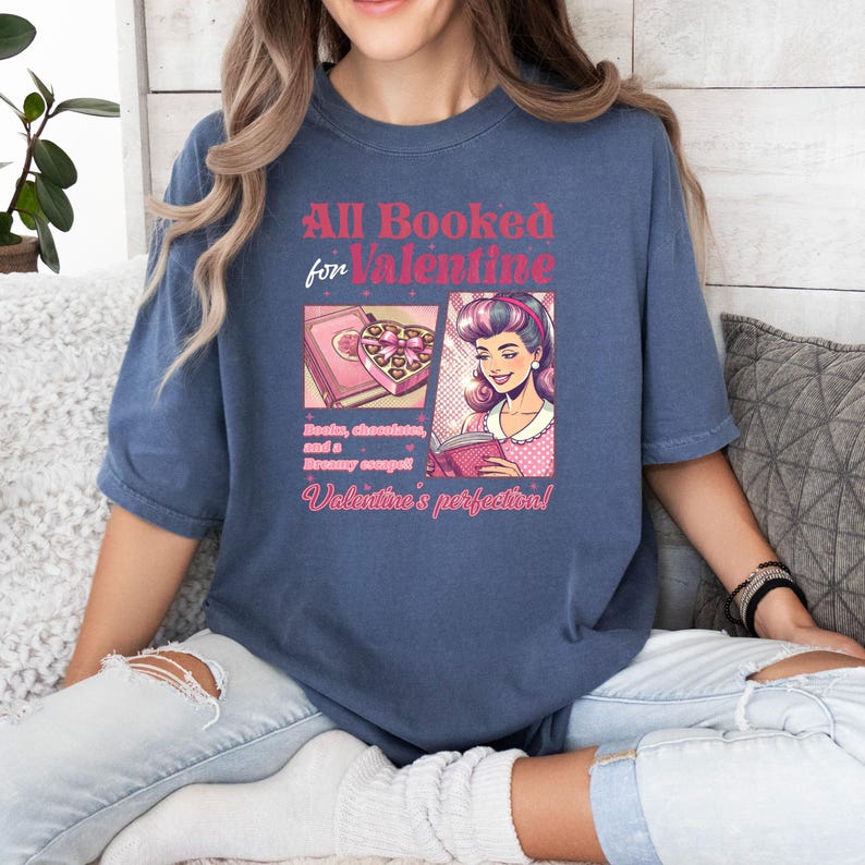 Puede incluir: Una camiseta azul con el texto "All Booked for Valentine" en rosa y blanco. La camiseta presenta una ilustraci&oacute;n de dibujos animados de una mujer leyendo un libro con una caja de chocolates en el fondo. El texto debajo de la ilustraci&oacute;n dice "Books, chocolates, and a dreamy escape! Valentine's perfection!"
