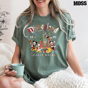 Puede incluir: Camiseta verde con un logotipo vintage de Disneyland con Mickey Mouse, Minnie Mouse, Donald Duck, Goofy y Pluto frente al Castillo de la Bella Durmiente. El texto dice "Disneyland Est. 1955 California".