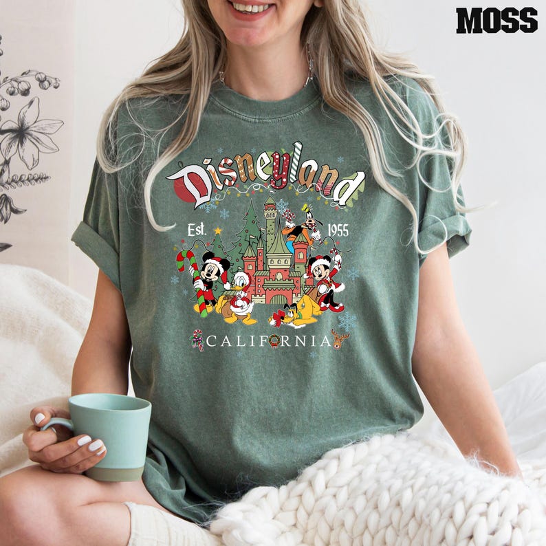 Puede incluir: Camiseta verde con un logotipo vintage de Disneyland con Mickey Mouse, Minnie Mouse, Donald Duck, Goofy y Pluto frente al Castillo de la Bella Durmiente. El texto dice "Disneyland Est. 1955 California".