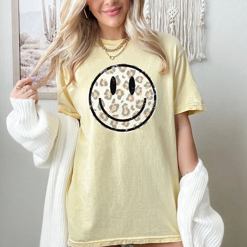 May include: A yellow t-shirt with a black and white smiley face graphic. The smiley face is filled with a leopard print pattern.