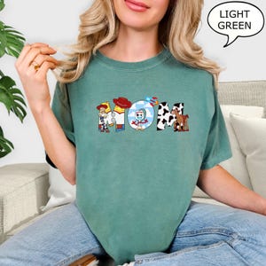 May include: Light green t-shirt featuring the word "MOM" in a colourful, cartoon design. Each letter is styled with a different character from a popular animated film. The shirt is made of a soft material.