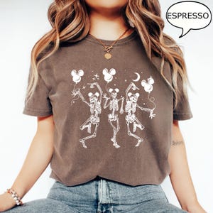 May include: Espresso-colored t-shirt featuring a white graphic of three dancing skeletons holding Mickey Mouse-shaped balloons. The design includes a crescent moon and stars. The word "ESPRESSO" is in a speech bubble.