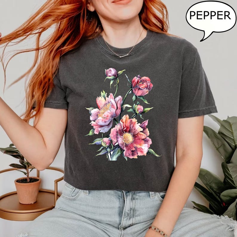 Pu&ograve; includere: T-shirt grigio scuro con un design floreale ad acquerello di peonie rosa e rosse e foglie verdi. La parola "PEPPER" &egrave; in una nuvoletta. La maglietta &egrave; in stile casual.