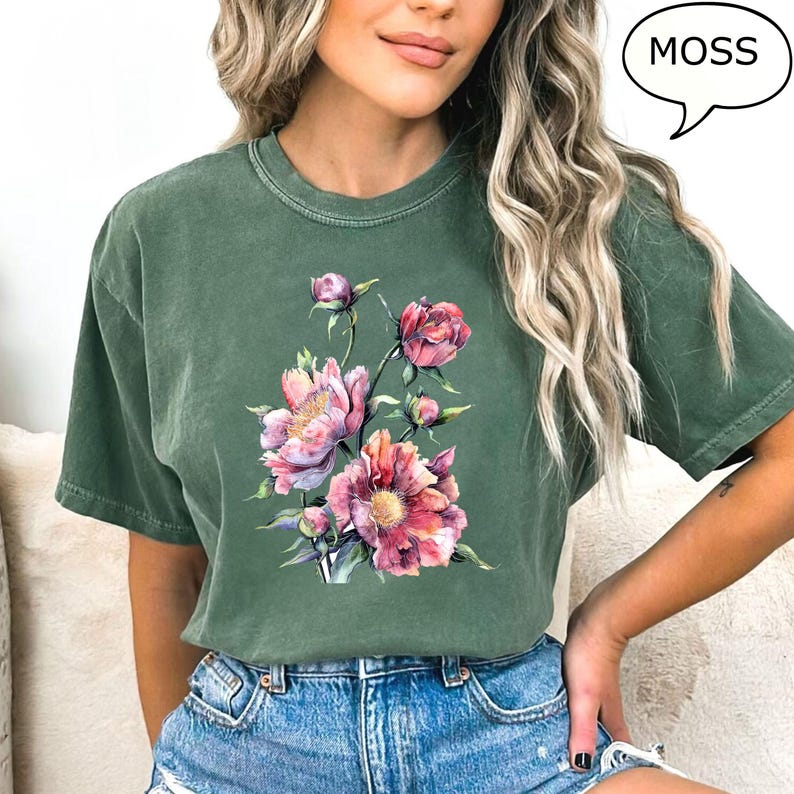 Pu&ograve; includere: T-shirt verde muschio con un design floreale ad acquerello di peonie rosa e rosse. La maglietta ha una vestibilit&agrave; ampia e maniche corte. La parola "MOSS" &egrave; in un fumetto.