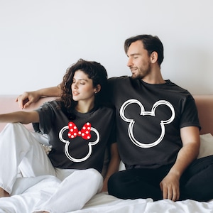May include: A couple wearing matching black t-shirts with white outlines of Mickey Mouse and Minnie Mouse heads. The Minnie Mouse shirt has a red bow with white polka dots.
