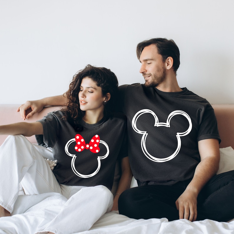 May include: A couple wearing matching black t-shirts with white outlines of Mickey Mouse and Minnie Mouse heads. The Minnie Mouse shirt has a red bow with white polka dots.