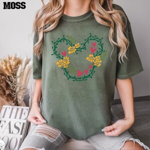 K&ouml;nnte beinhalten: Ein gr&uuml;nes T-Shirt mit einem floralen Mickey-Mouse-Design. Das Design enth&auml;lt gelbe Blumen, gr&uuml;ne Bl&auml;tter und rote Herzen.