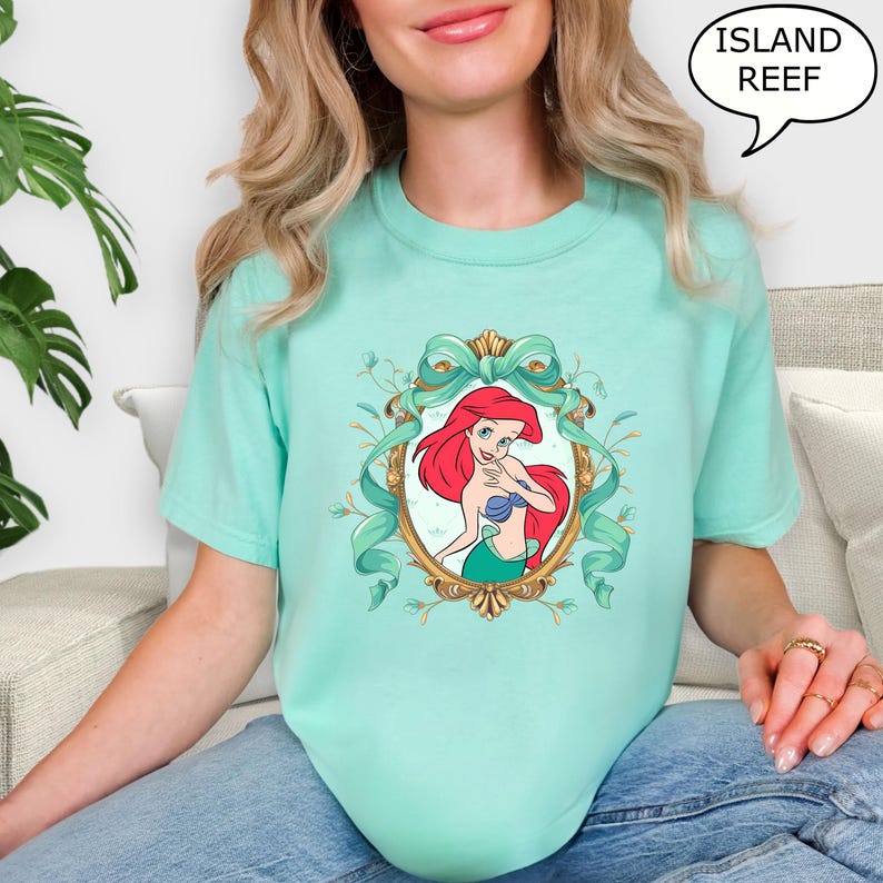 May include: A light teal t-shirt featuring a framed illustration of a red-haired mermaid with a blue seashell top and green tail. The frame is gold with teal ribbons and floral accents. The text "ISLAND REEF" is in a speech bubble.