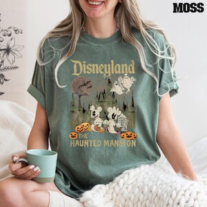 May include: A green t-shirt with a vintage-style graphic of Mickey Mouse and a ghost in front of the Haunted Mansion at Disneyland. The text "Disneyland" and "The Haunted Mansion" are printed on the shirt.