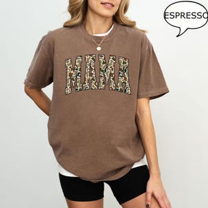May include: A brown t-shirt with a large "MAMA" graphic printed on the front. The letters are filled with a camouflage pattern.