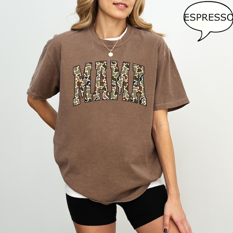 May include: A brown t-shirt with a large "MAMA" graphic printed on the front. The letters are filled with a camouflage pattern.
