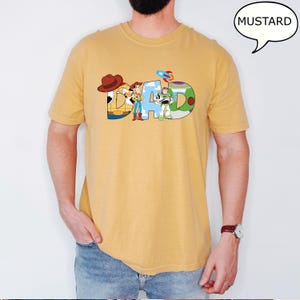 May include: Mustard-coloured t-shirt featuring the word "DAD" in a colourful, cartoon-style font. The letters are designed with characters from a popular animated film. The shirt is a solid colour and has short sleeves.
