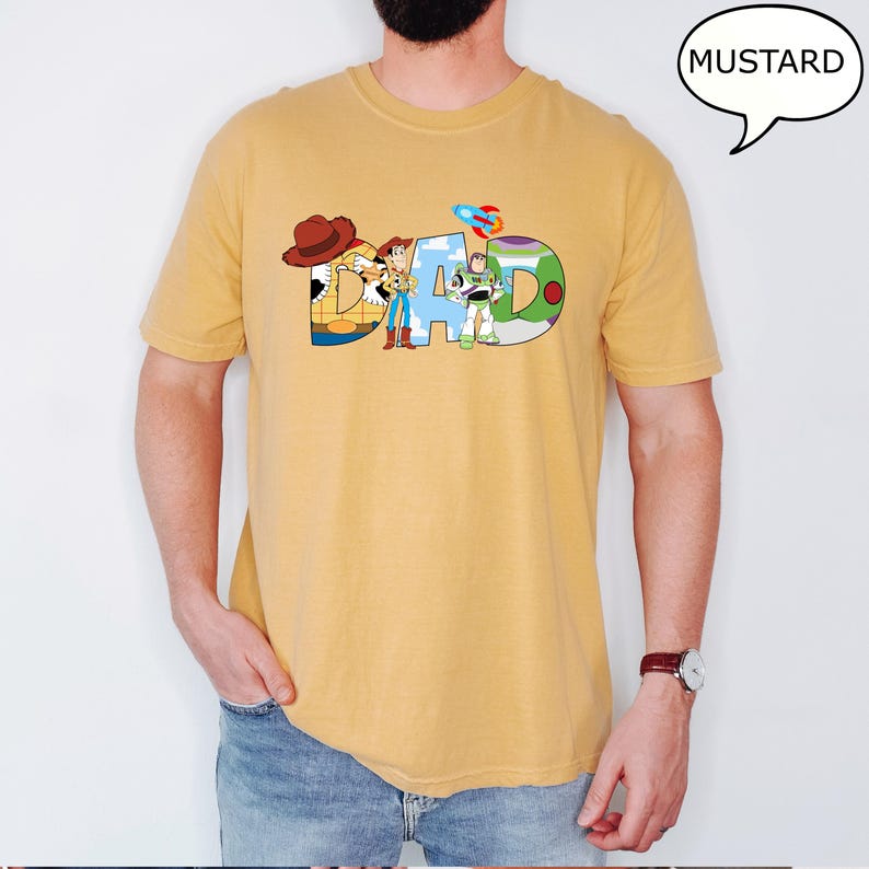 May include: Mustard-coloured t-shirt featuring the word "DAD" in a colourful, cartoon-style font. The letters are designed with characters from a popular animated film. The shirt is a solid colour and has short sleeves.