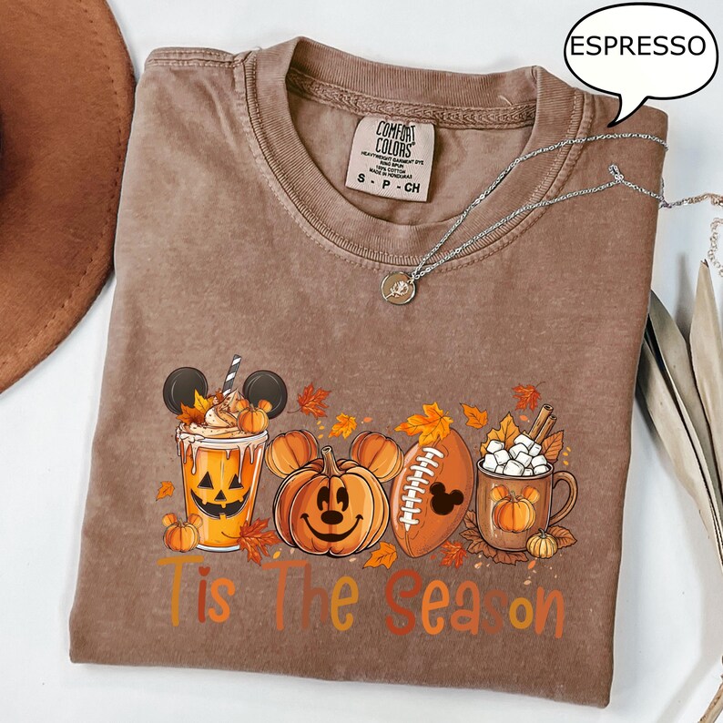 May include: Espresso-colored Comfort Colors t-shirt with a fall-themed graphic. The design includes a pumpkin spice drink, jack-o'-lanterns, a football, and a mug of hot cocoa. The text "Tis The Season" is printed on the front.