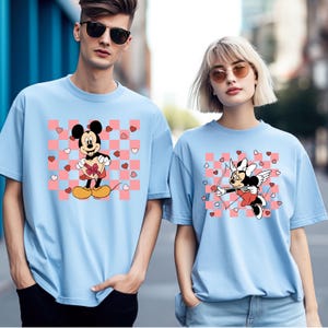 Puede incluir: Camisetas de color azul claro con dise&ntilde;os de Mickey y Minnie Mouse. Una camiseta muestra a Mickey sosteniendo un regalo en forma de coraz&oacute;n, la otra a Minnie como Cupido con arco y flecha. Ambas camisetas tienen un fondo a cuadros rosa y blanco con corazones.
