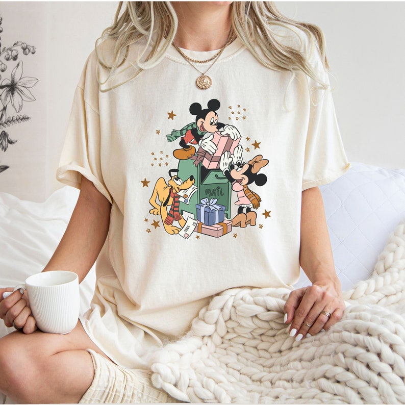 May include: A cream coloured t-shirt featuring a vintage cartoon graphic of Mickey Mouse, Minnie Mouse, and Pluto standing by a mailbox with presents and stars. The text "Mail" is on the mailbox.