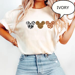 Pu&ograve; includere: T-shirt color avorio con tre sagome della testa di Topolino con disegni animalier: zebra, leopardo e giraffa. La parola "IVORY" &egrave; in una nuvoletta. La maglietta &egrave; abbinata a jeans blu.