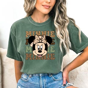 May include: A green short-sleeved t-shirt with a graphic print of Minnie Mouse wearing a safari hat and a leopard print bow. The text "Minnie Mouse Minnie" is printed in a leopard print font.