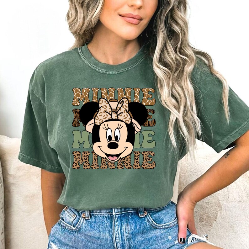 May include: A green short-sleeved t-shirt with a graphic print of Minnie Mouse wearing a safari hat and a leopard print bow. The text "Minnie Mouse Minnie" is printed in a leopard print font.