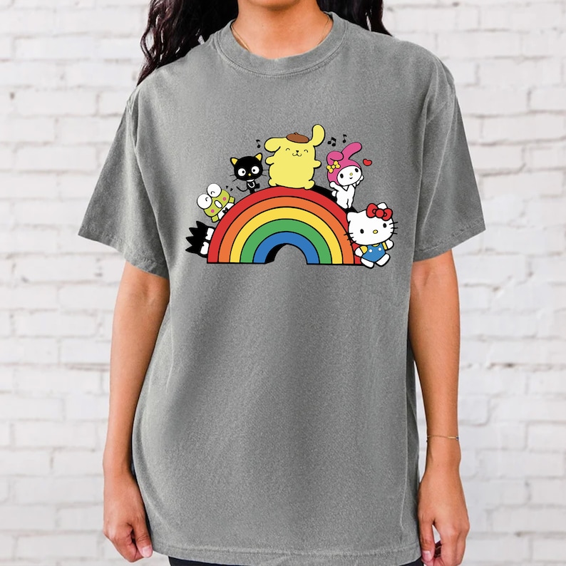 Pode incluir: Uma camiseta cinza com um arco-&iacute;ris e personagens de desenhos animados. Os personagens incluem Hello Kitty, My Melody, Keroppi e um cachorro amarelo.