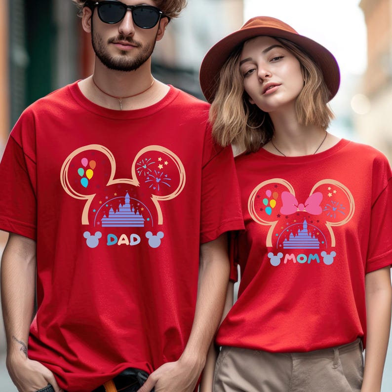 Pode incluir: Duas camisetas vermelhas com um design de silhueta do Mickey Mouse. As camisetas apresentam um castelo de desenho animado, bal&otilde;es e fogos de artif&iacute;cio. O texto "DAD" e "MOM" est&aacute; impresso nas camisetas.