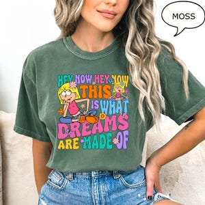 Może przedstawiać: Zielona koszulka z kr&oacute;tkim rękawem z kolorowym nadrukiem graficznym, na kt&oacute;rym widnieje napis "Hey Now Hey Now This Is What Dreams Are Made Of". Grafika przedstawia dwie kresk&oacute;wkowe dziewczyny i kwiaty.