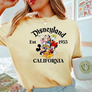 Puede incluir: Camiseta amarilla con un gr&aacute;fico negro de Mickey Mouse y sus amigos. El gr&aacute;fico dice "Disneyland Est 1955 California".