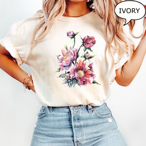 Pu&ograve; includere: T-shirt color avorio con un design floreale ad acquerello con peonie rosa e viola e foglie verdi. La parola "IVORY" &egrave; in una nuvoletta. La maglietta ha maniche corte e una vestibilit&agrave; ampia.