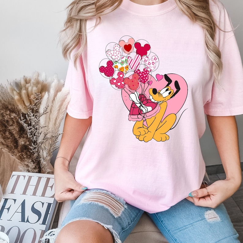 Może przedstawiać: R&oacute;żowy T-shirt z kresk&oacute;wkowym psem trzymającym pudełko czekoladek w kształcie serca. Pies jest otoczony r&oacute;żowymi i czerwonymi balonami w kształcie serca.