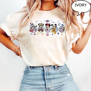 May include: Ivory-colored t-shirt featuring a nautical design with cartoon characters. The design includes Mickey Mouse, Minnie Mouse, Goofy, Pluto, and Donald Duck, each in a circular frame with nautical elements. The word "IVORY" is in a speech bubble.