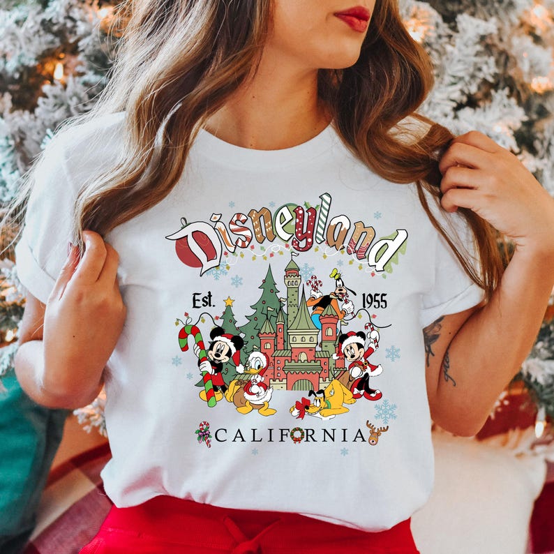 Puede incluir: Camiseta blanca con un gr&aacute;fico de Disneyland con tem&aacute;tica navide&ntilde;a. El gr&aacute;fico presenta a Mickey Mouse, Donald Duck, Goofy y Pluto frente al castillo de la Bella Durmiente. El texto "Disneyland Est. 1955 California" est&aacute; impreso en la camiseta.