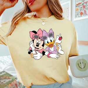 Puede incluir: Una camiseta amarilla con un estampado gr&aacute;fico de Minnie Mouse y Daisy Duck con gafas de sol y sosteniendo conos de helado. El cono de helado tiene una fresa en la parte superior.