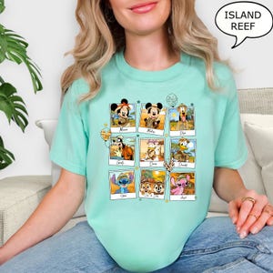 Puede incluir: Una camiseta turquesa con un gr&aacute;fico de personajes de Disney en un entorno de safari. Los personajes incluyen Mickey Mouse, Minnie Mouse, Pluto, Goofy, Donald Duck, Daisy Duck, Stitch, Chip y Dale. El texto "Island Reef" est&aacute; en un bocadillo de di&aacute;logo en la esquina superior derecha de la imagen.