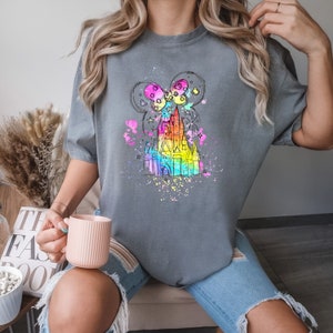 Puede incluir: Una camiseta gris con un dise&ntilde;o de acuarela arco&iacute;ris de un castillo y orejas de Mickey Mouse. El dise&ntilde;o est&aacute; rodeado de salpicaduras de pintura blanca.