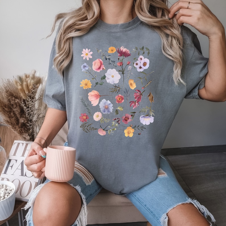 May include: A gray t-shirt with a floral print featuring a variety of flowers in shades of pink, purple, yellow, and red.