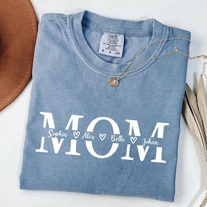 May include: A blue Comfort Colors t-shirt with the word "MOM" in large white letters. Below the word "MOM" are the names Sophia, Alex, Belle, and Johan, each with a heart symbol. A gold necklace with a pendant is on the shirt.