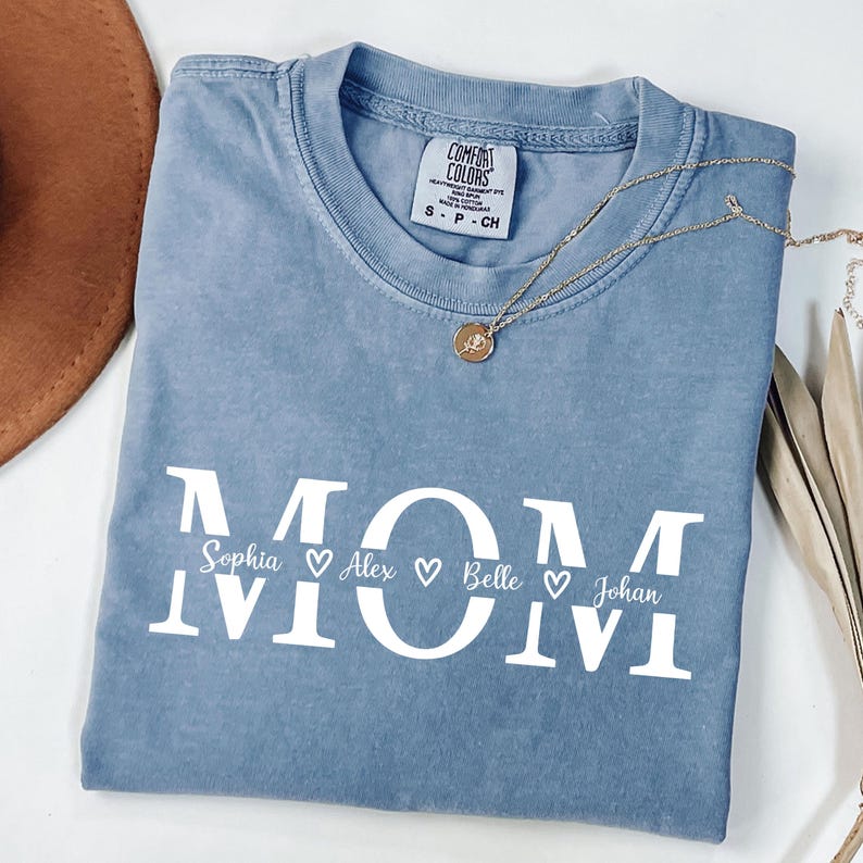 May include: A blue Comfort Colors t-shirt with the word "MOM" in large white letters. Below the word "MOM" are the names Sophia, Alex, Belle, and Johan, each with a heart symbol. A gold necklace with a pendant is on the shirt.