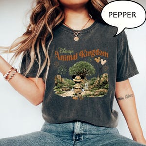 May include: Dark gray t-shirt featuring the text "Disney's Animal Kingdom" in orange and green. The graphic includes Mickey Mouse in safari attire, a tree, and a landscape scene. A speech bubble with the word "PEPPER" is visible.
