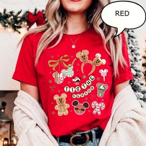 May include: A red t-shirt with a festive design featuring gingerbread men, Mickey Mouse-shaped ornaments, candy canes, and the phrase "'Tis the Season." The shirt has short sleeves.