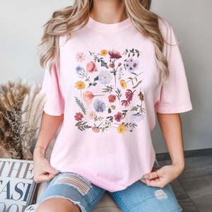 May include: A pink t-shirt with a floral print featuring a variety of flowers in shades of pink, blue, yellow, and red.