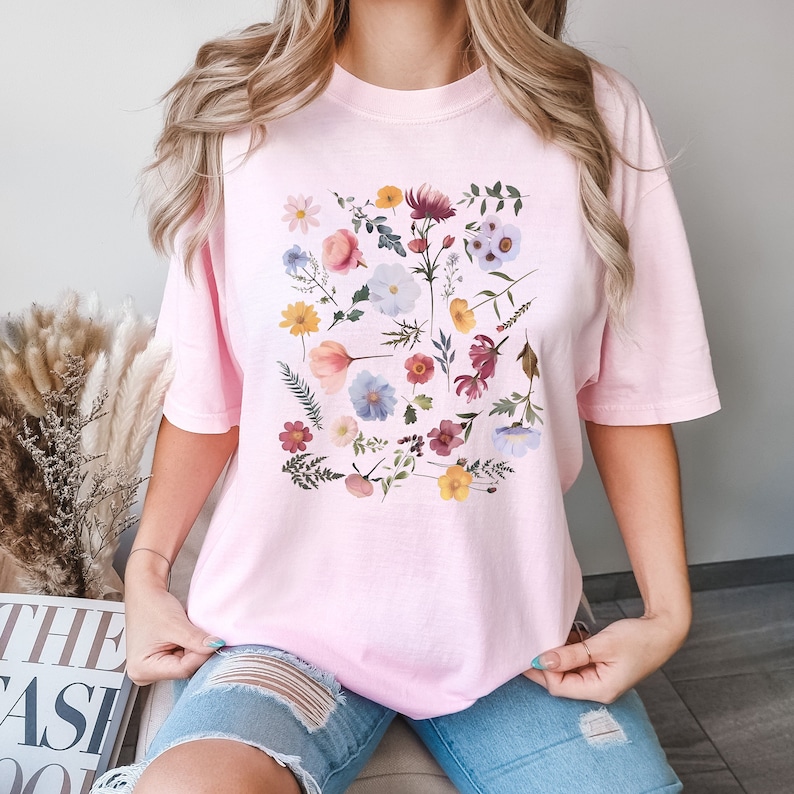 May include: A pink t-shirt with a floral print featuring a variety of flowers in shades of pink, blue, yellow, and red.