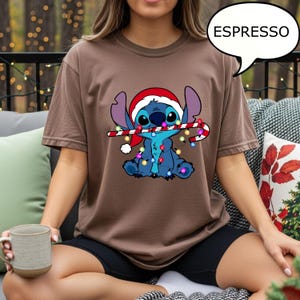 Puede incluir: Una camiseta marr&oacute;n con un personaje de dibujos animados que lleva un gorro de Pap&aacute; Noel y sostiene un bast&oacute;n de caramelo con luces navide&ntilde;as. La palabra "ESPRESSO" est&aacute; en un bocadillo. La camiseta es de color liso.