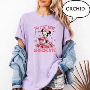 Puede incluir: Camiseta morada orqu&iacute;dea con un gr&aacute;fico de Minnie Mouse. El gr&aacute;fico incluye el texto "I'M JUST HERE for the CHOCOLATE" con corazones y un personaje de Minnie Mouse sosteniendo una caja de regalo. La camiseta tiene mangas enrolladas y un ajuste relajado.