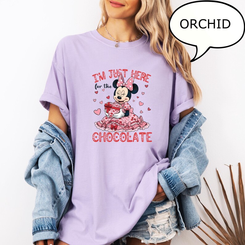 Puede incluir: Camiseta morada orqu&iacute;dea con un gr&aacute;fico de Minnie Mouse. El gr&aacute;fico incluye el texto "I'M JUST HERE for the CHOCOLATE" con corazones y un personaje de Minnie Mouse sosteniendo una caja de regalo. La camiseta tiene mangas enrolladas y un ajuste relajado.