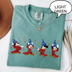 May include: Light green t-shirt with a graphic of Mickey Mouse in a wizard costume. The design features Mickey in a red robe and blue wizard hat, with yellow stars. The shirt has a Comfort Colors tag.