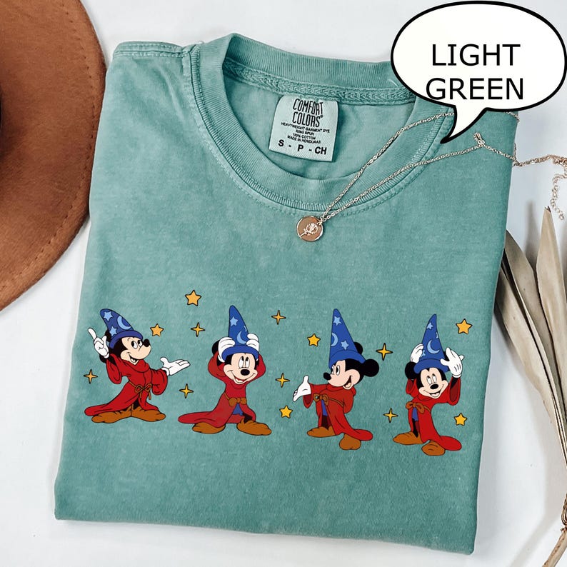 May include: Light green t-shirt with a graphic of Mickey Mouse in a wizard costume. The design features Mickey in a red robe and blue wizard hat, with yellow stars. The shirt has a Comfort Colors tag.