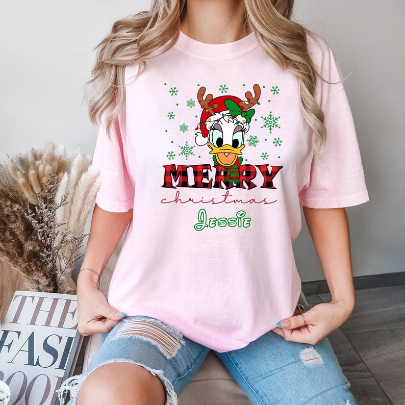 Op de afbeelding: Roze T-shirt met een cartoon afbeelding van een eend die een kerstmanmuts en rendiergewei draagt. De tekst "Merry Christmas Jessie" staat op het T-shirt gedrukt.